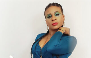 Posing in front of a cream background, Lachi, a Black woman with long, black, braided hair and shimmering green eyeshadow. She is wearing a teal dress and gold necklace. In her hand, she is holding her gold-coloured cane, which is folded-up.