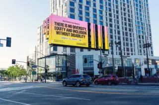 Photo of a billboard of bold text over a bright pink, orange and yellow gradient background that reads &ldquo;There&rsquo;s no diversity, equity, and inclusion without disability.&rdquo; The billboard hangs on a tall building overlooking a wide street.