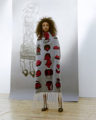 Model wearing a grey fringed garment with red and black abstract face designs, standing in front of a large sketch of a woman in a patterned dress