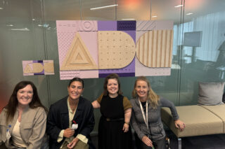 Orla, Sinead and Laia, members of the Tilting the Lens team, smile for a photo with a member of the Google Accessibility Discovery Centre (ADC) team. They are at the ADC in Dublin crouched under the tactile ADC sign.