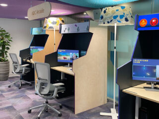 The image shows the ADC Arcade with three individual game setups, each with its own desk, monitor and gaming chair. Each station highlights different accessibility solutions. Positioned between the setups are two floor lamps with brightly coloured lampshades, adding a vibrant and playful element to the environment.