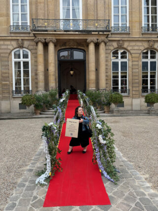 Image Two: Sin&eacute;ad Burke, a white Little Person stands on the red carpet ramp leading up to the British Ambassador&rsquo;s Residence in Paris. She is holding a sign which reads &ldquo;Tilting the Lens, Services: People and Culture, Built Environment, Product and Service Design, Communication and Representation&rdquo;.