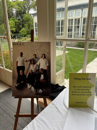 A large photo of the Disabled art collective Trifle Studio taken from the May 2023 issue of British Vogue sits proudly on a wooden easel. On a table to the side, a sign reads &ldquo;Tilting the Lens, we craft solutions to intentionality build an accessible and equitable world&rdquo;, next to it the Braille issue of the magazine is positioned.