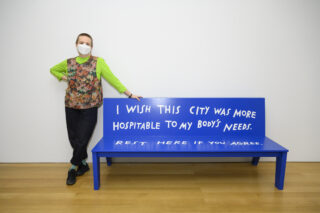 Finnegan Shannon, posed next to a bright blue bench. Their handwriting on the bench reads, "I wish this city was more hospitable to my body's needs. Rest here if you agree." Finnegan is a boyish white person with a buzz cut. They are wearing a KF94 mask, a fluorescent lime shirt, a floral fisherman-style vest.