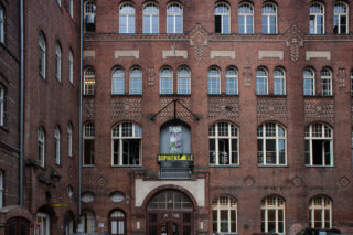 The facade of the Sophiens&aelig;le, as viewed from the inner courtyard by Joe Goergen. It is a multi-story brick building with the logo of Sophiensaele in a black lightbox that is much more modern and brings some contrast to the entrance. Above the lightbox is a small poster for &ldquo;Lucky Trimmer&rdquo;.