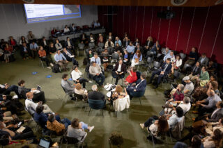 An overview of the room that the session took place in. It is large with the 9 speakers arranged in a circle on chairs in the centre. The audience are seated around them in concentric circles. There is a screen on one wall with information about the session.