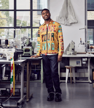 Artist Christian Ovonlen is a Black man wearing a unique garment&mdash;a jacket&mdash;created in collaboration with R&AElig;BURN. Christian stands in their Hackney studio in East London, surrounded by sewing machines, materials, and a large window in the background. He stands proudly, donning an orange jacket with red, black, and mint lettering bearing the words "CHANGE YOUR MIND." The textile, designed by Andre Williams and hand screen printed on R&AElig;BURN&rsquo;s signature parachute fabric, makes a bold statement. This phrase has become an emblematic call to action as the artist encourages audiences, creatives, commissioners, and brands to think differently about how they can foster greater inclusivity and access within their fields.