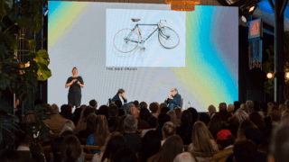 Two speakers on stage are in discussion with a large screen behind them. A sign language interpreter stands to one side facing the audience.