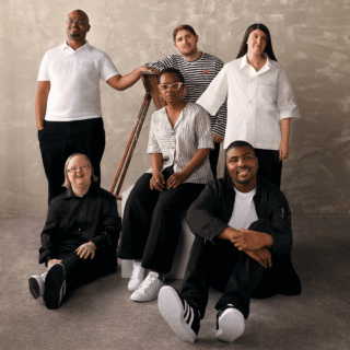 Top row, from the left: three artists, Andre Williams, a black man with glasses, is wearing a white top and black trousers. Stanley Galton, a white man with brown hair, is dressed in a striped black and white top. Nancy Clayton, a white woman with long brown hair, is wearing a white shirt and black trousers. Bottom row, from left: Three artists, including Lisa Trim, a white woman with glasses and blonde hair, sit on the floor in black shirts and trousers. Ntiense Eno-Amooquaye, a black woman with short black hair and pink-rimmed glasses, is sitting on a white block wearing a grey and white shirt, black trousers, and white trainers. Lastly, Christian Ovonlen, a black man with black hair, is sitting on the floor wearing a black shirt, a white T-shirt, black trousers, and Adidas trainers. They are all positioned against a grey backdrop.