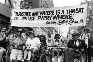 A black and white picture depicting Judy Heumann, a white power wheelchair user, amongst other Disabled demonstrators. A banner above their heads reads: &ldquo;Injustice anywhere is a threat to justice everywhere.&rdquo; &ndash; a quote by Martin Luther King Jr.